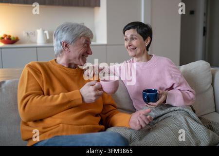 Un mari et une femme seniors souriants boivent du thé ensemble lors d'une conversation détendue sur le canapé de la maison Banque D'Images