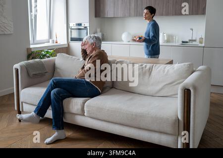 Mari âgé frustré assis sur le canapé dans la pièce, écoutant tandis que son épouse se plaint et argumente à l'arrière-plan. Banque D'Images