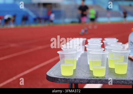 Gobelets en plastique transparent remplis d'une boisson électrolytique jaune-vert sur une table à côté d'une piste de course rouge, des coureurs flous s'entraînent en arrière-plan Banque D'Images