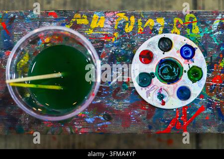 Palette d'aquarelle ronde avec des couleurs de peinture mélangées à côté d'une tasse d'eau de rinçage verte contenant deux pinceaux, sur une table tachée de peinture. Banque D'Images
