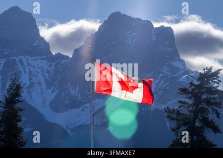 Drapeau national canadien flottant sur un mât sous un soleil éclatant, avec des reflets de lumière et des arbres à feuilles persistantes au premier plan devant les Three Sist enneigés et emblématiques Banque D'Images