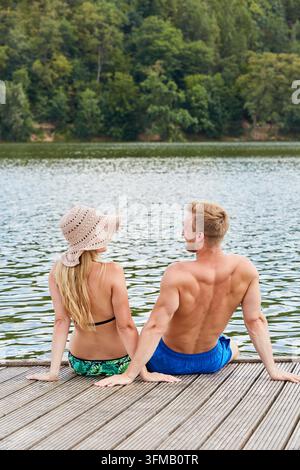 Jeune couple assis sur une crête en bois d'un lac de baignade en vacances en été Banque D'Images