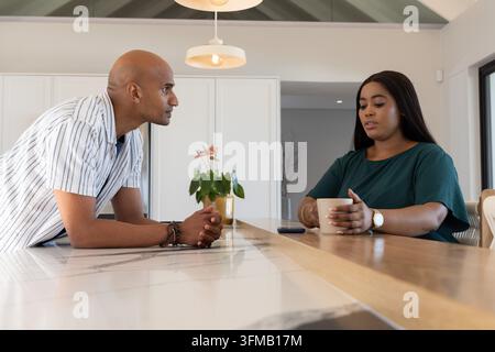 Couple homme indien et femme afro-américaine discutant dans la cuisine, tenant une tasse, smartphone à proximité. Moderne, minimaliste, intérieur, maison, concept ouvert, îlot, suspension Banque D'Images
