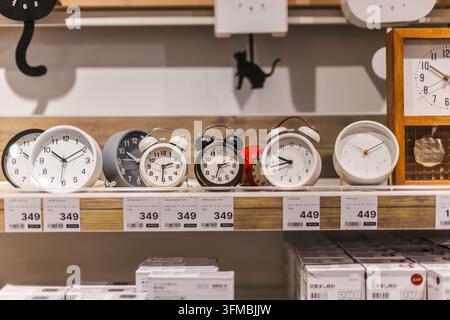 Bangkok, Thaïlande, 4 mai 2026 : Une étagère de magasin présente divers réveils analogiques et horloges murales en blanc, noir et rouge. Banque D'Images