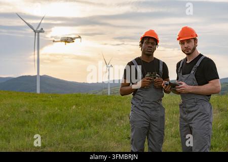 Deux ingénieurs casqués utilisant un drone et des jumelles dans un parc éolien Banque D'Images