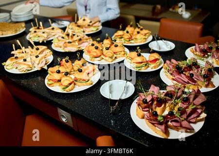 Assortiment d'apéritifs disposés sur une table de buffet noire pour le catering, comprenant des canapés, des sandwichs doigts, des olives, du fromage, des cornichons et une garniture d'herbes Banque D'Images