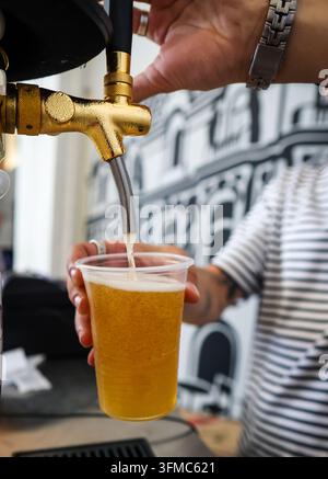 Un barman verse une bière fraîche de la pression d'un robinet doré étincelant dans un gobelet en plastique. Banque D'Images