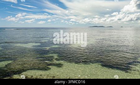 Les eaux turquoise claires reflètent le ciel, avec une île tranquille à l'horizon. Seychelles. Banque D'Images