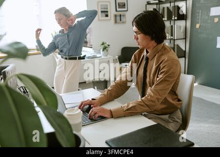 Homme adulte travaillant sur un ordinateur portable pendant qu'une femme danse au bureau Banque D'Images
