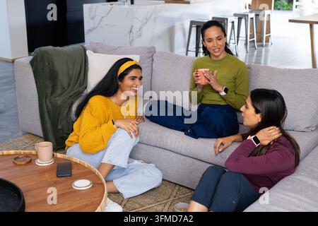 Différentes amies féminines discutant et sirotant des tasses sur un canapé gris à la maison avec un téléphone sur la table Banque D'Images
