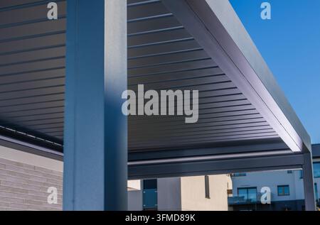 Détail architectural d'une pergola métallique sombre et d'un toit à lamelles à côté d'une maison moderne sous un ciel bleu clair. Banque D'Images