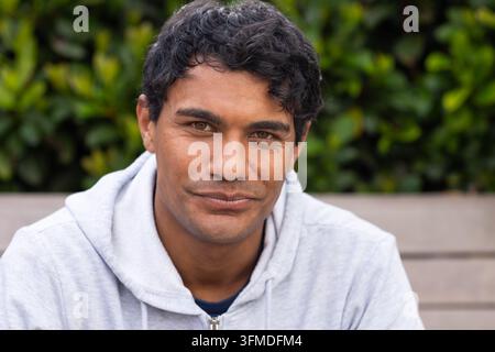 Homme afro-américain assis sur un banc près d'une haie, portant un sweat à capuche gris et un t-shirt à col rond sombre Banque D'Images
