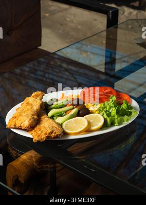 Du poisson frit doré repose à côté de salades vertes fraîches. Des tomates vives et des tranches de citron ajoutent de la couleur et de la fraîcheur. De la laitue abondante et des carottes complètent la vivacité Banque D'Images