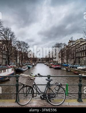 Un vélo classique avec un panier en osier repose sur la rambarde d'un pont surplombant un canal historique bordé de péniches à Amsterdam sous un ciel menaçant. Banque D'Images