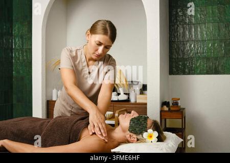 Femme d'âge moyen allongée sur une table de massage dans un spa. La massothérapeute travaille sur ses épaules avec une attention concentrée. La pièce présente un décor naturel et un éclairage doux Banque D'Images