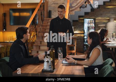 Serveur en uniforme noir prenant les commandes d'un couple interethnique élégant lors d'un dîner romantique dans un restaurant de luxe. Banque D'Images
