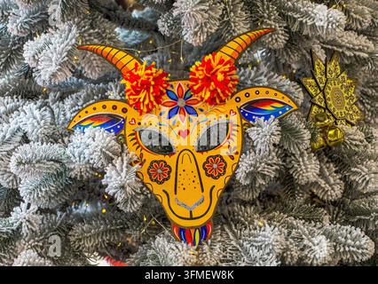 Masque de carnaval vibrant et décoratif en forme de chèvre, doté de cornes et de pompons, orné d'un motif floral peint, posé sur un fond de branches d'épicéa Banque D'Images
