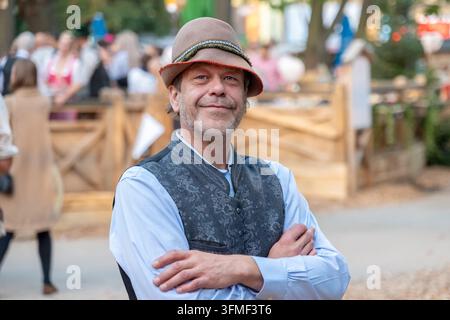 Un homme souriant portant un chapeau se tient les bras croisés lors d'un festival en plein air. Banque D'Images