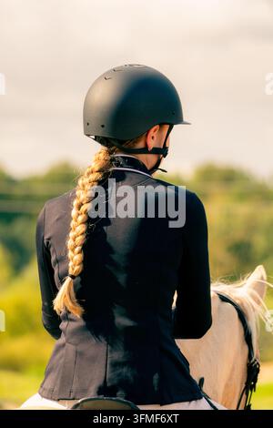 Cavalière avec une longue tresse blonde portant un casque d'équitation et assise sur un cheval en plein air Banque D'Images