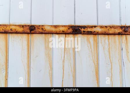 Photographie de texture en gros plan montrant une sangle de charnière en fer fortement rouillée avec des gouttes de corrosion coulant le long de planches de bois peintes en blanc verticales. Banque D'Images