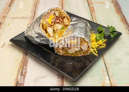 Moitiés de burrito enveloppées dans du papier aluminium avec une garniture au poulet, œuf et bacon, servies avec des pommes de terre coupées en julienne et du persil sur une assiette noire. Banque D'Images