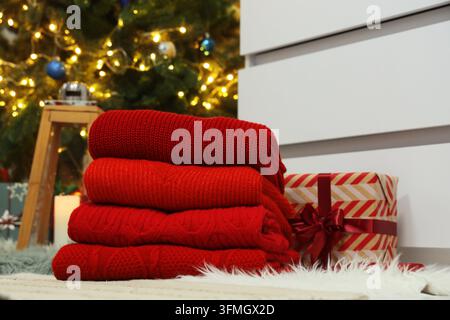 Préparation des fêtes de Noël, pile de pulls tricotés rouges avec boîte cadeau et guirlandes lumineuses festives dans un intérieur confortable pour les achats de la saison hivernale Banque D'Images