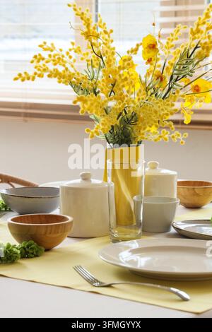 Décoration printanière pour la maison et le dîner, fleurs de mimosa jaunes dans un vase avec de la vaisselle en céramique sur la table pour le petit-déjeuner ou le brunch dans une pièce lumineuse baignée de soleil Banque D'Images