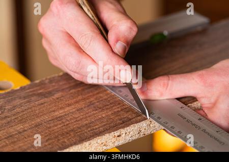 Close-up de mains d'artisans utilisant un couteau de marquage et une règle en métal sur une planche de bois. Concept de travail du bois, mesure précise, menuiserie et artisanat Banque D'Images