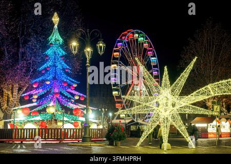 Marché de Noël la nuit avec une installation géante en forme d'étoile lumineuse, un sapin de Noël néon lumineux et une grande roue illuminée à Asti, Piémont, Italie Banque D'Images