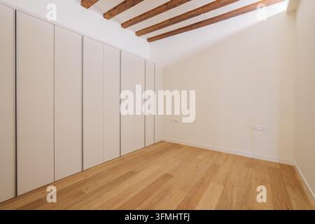 Pièce minimaliste vide avec un sol en chêne clair, des murs blancs, des poutres apparentes au plafond et des armoires encastrées sur toute la hauteur avec portes affleurantes Banque D'Images