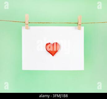 Composition minimale avec une forme de cœur rouge sur une carte blanche vierge accrochée à une corde sur un fond vert, symbolisant l'amour, avec espace de copie. Banque D'Images