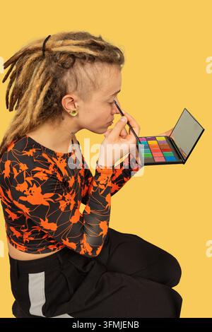 Jeune personne aux dreadlocks appliquant du fard à paupières depuis une palette colorée sur un fond jaune vif Banque D'Images