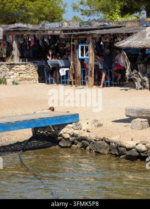 Pirovac, Croatie, 16 août 2026 Bar de bord de mer détendue avec toit de paille, tables en bois, visiteurs assis, mur en pierre, sol sablonneux et eau claire et peu profonde Banque D'Images