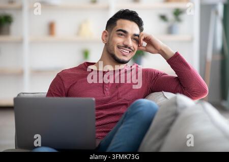 Rêveur arabe se reposant sur un canapé à la maison, en utilisant un ordinateur portable et souriant, à la recherche d'un emploi en ligne, espace de copie. Heureux jeune homme du moyen-Orient surfant sur dati Banque D'Images