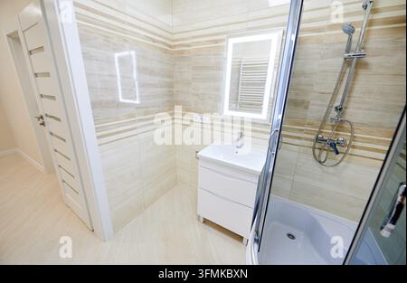 Intérieur de salle de bain moderne avec cabine de douche, lavabo avec meuble de rangement et miroir éclairé. Salle d'eau lumineuse avec carrelage à rayures beiges, porte en verre, accessoires chromés et vue depuis la porte Banque D'Images