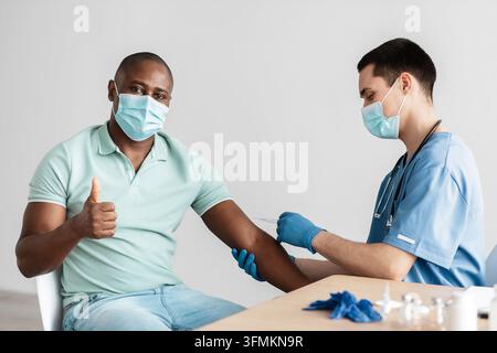L'homme positif obtient le vaccin, pouce vers le haut, faisant excellent et signe d'approbation geste. Jeune médecin dans le masque de protection fait l'injection de l'adulte africain ameri Banque D'Images
