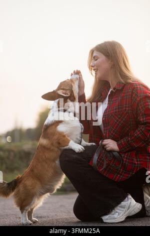 Jeune femme marchant avec son chien Pembroke Welsh Corgi dans un parc verdoyant par une journée ensoleillée. La femme et son animal de compagnie profitant d'une promenade en plein air entourée d'arbres a Banque D'Images