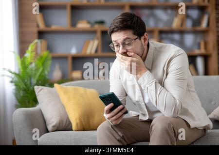 Jeune homme assis sur un canapé dans son salon, regardant un smartphone avec une expression choquée et inquiète, se couvrant la bouche d'incrédulité après avoir lu de mauvaises nouvelles en ligne Banque D'Images