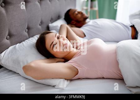 Bonne jeune femme caucasienne se reposant au lit à côté de son mari noir à la maison, couchée avec les mains derrière sa tête, se sentant détendue le dimanche matin. Marr Banque D'Images
