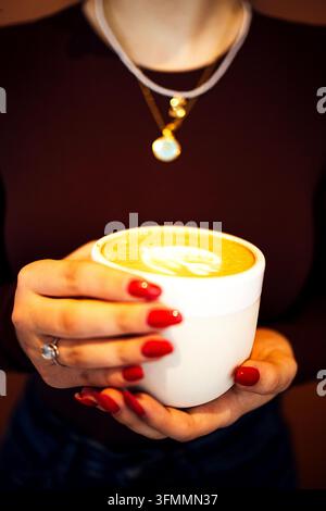 Femme aux ongles manucurés en rouge tenant une tasse de latte blanche sous des colliers superposés, tandis qu'un art en mousse en forme de cœur est visible contre des vêtements sombres Banque D'Images