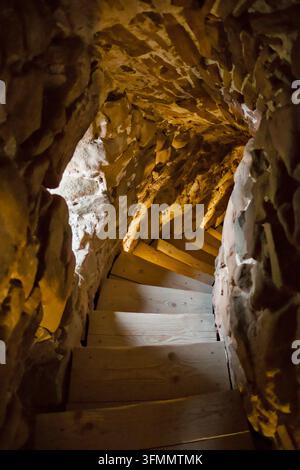 Regard vers le bas d'un escalier en bois étroit et sinueux intégré dans un tunnel de tour en pierre brute, éclairé par une lumière ambiante intérieure chaude. Banque D'Images