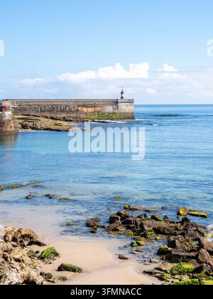 Pier de pierre avec phare par temps ensoleillé – Un mur de port historique en pierre avec un petit phare s’étend dans la mer bleue et calme à côté d’une plage rocheuse couverte de mousse Banque D'Images