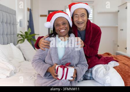 Homme asiatique âgé et femme africaine assis dans la chambre portant des chapeaux de Noël et tenant une boîte cadeau Noël Banque D'Images