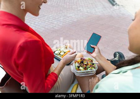 Deux femmes d'affaires diverses se détendent en plein air après le travail, profitant de salades à emporter saines tandis qu'une vérifie son smartphone pendant une pause déjeuner informelle Banque D'Images