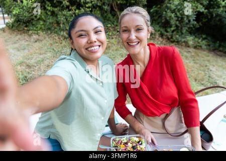 Deux femmes d'affaires souriantes prennent un selfie à une table extérieure pendant une pause déjeuner après le travail, profitant d'une salade fraîche et d'une conversation amicale dans un gr Banque D'Images
