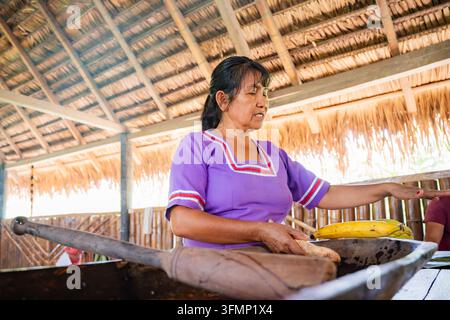 Femme amazonienne en robe violette traditionnelle préparant une nourriture ancestrale avec des banains dans une cuisine rustique en paille, préservant la culture culinaire indigène Banque D'Images