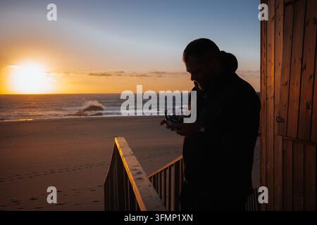 Homme en silhouette opérant une télécommande de drone depuis une structure en bois au bord de la plage au coucher du soleil, surplombant les dunes de sable et l'océan Atlantique sur le Portug Banque D'Images