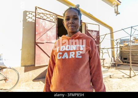 Une jeune femme portant un sweat à capuche orange se tient à côté de son vélo devant sa maison à porte métallique, Vakinankaratra, Antsirabe, Madagascar Banque D'Images