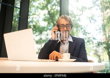 Un homme d'affaires senior parle au téléphone tout en travaillant sur un ordinateur portable dans un bureau lumineux. Il est assis à un bureau avec du café, examinant le travail pendant un professeur Banque D'Images