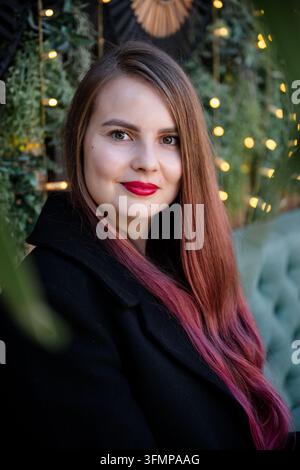 Portrait d'une femme souriante aux cheveux bruns longs et pointes roses, au rouge à lèvres rouge et au manteau noir, posée à côté d'un sapin de Noël décoré à la lueur chaude Banque D'Images